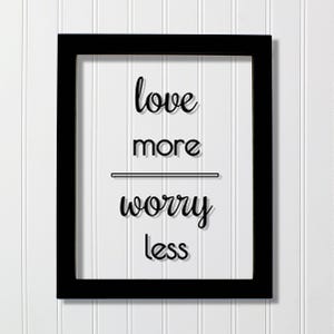 Op de afbeelding: Een rechthoekige zwarte lijst met een wit bord met de zwarte tekst "love more" boven een horizontale lijn, gevolgd door de tekst "worry less" onder de lijn. De belettering gebruikt een combinatie van script- en schreefloze lettertypes.