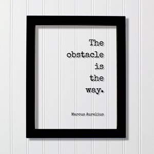 Puede incluir: Un marco de fotos rectangular negro que contiene una impresión blanca con el texto negro estilo máquina de escribir: The obstacle is the way. Marcus Aurelius. El texto está dispuesto en cuatro líneas, con el nombre del autor en la esquina inferior derecha.