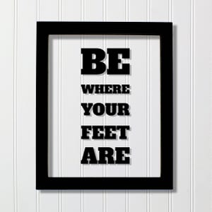 Op de afbeelding: Een rechthoekige zwarte lijst met een witte achtergrond en de zwarte tekst BE WHERE YOUR FEET ARE gedrukt in een vetgedrukt bloklettertype. De tekst is gecentreerd in de lijst, wat zorgt voor een duidelijke en leesbare boodschap.