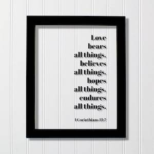 May include: A rectangular black frame containing a clear pane with black text that reads: Love bears all things, believes all things, hopes all things, endures all things. 1 Corinthians 13:7. The lettering is centered and features a drop shadow.