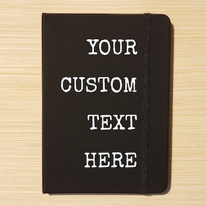 May include: Black notebook with a black elastic closure. The notebook has white text that reads "YOUR CUSTOM TEXT HERE".