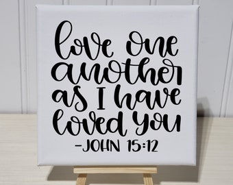 May include: White canvas with black lettering that reads "love one another as I have loved you - JOHN 15:12". The canvas is on a small wooden easel.