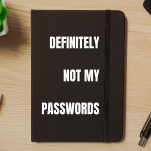May include: Black notebook with the text "DEFINITELY NOT MY PASSWORDS" in white. The notebook has an elastic band closure and is lying on a wooden surface. A pen and a small plant are also visible.