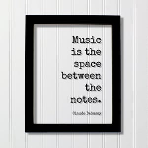 May include: A black rectangular frame containing a white print with the black typewriter-style text: Music is the space between the notes. Claude Debussy. The lettering features a subtle drop shadow effect against the plain white background.