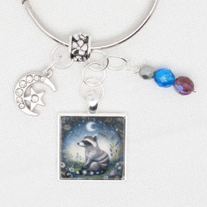 May include: A silver charm bracelet with a square charm featuring a raccoon sitting in a field of flowers under a crescent moon. The bracelet also has a silver charm in the shape of a crescent moon with a star, a silver bead with flower designs, and three beads in blue and purple.