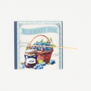 May include: A square, light blue sign with the words "BLUEBERRY JAM" in a banner. The sign features a jar of jam, a basket of blueberries, and the text "Since 1899". A gold-colored stick is placed across the sign.