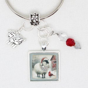 May include: A silver-toned charm bracelet with a sheep charm, a square pendant with a winter scene of a sheep and a red bird, and a red and clear bead charm. The pendant has a silver frame and a winter scene.