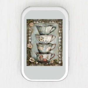 May include: A decorative tin featuring a stack of four antique teacups. The teacups are white with floral designs and gold trim. The background is a blue and gray plaid pattern with rose accents.