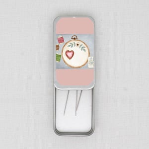 Pode incluir: Uma caixa de agulhas retangular de metal com tampa rosa com um desenho em aquarela de um bordado em forma de coração em um bastidor, carretéis de linha e agulhas. A caixa aberta revela três agulhas.