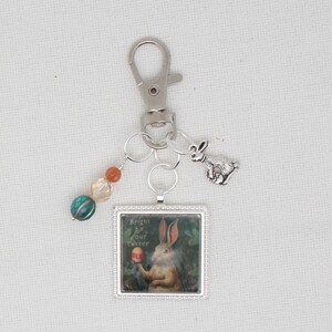 May include: A silver keychain with a square pendant featuring a rabbit holding an egg, with the text "Bright be your Easter." It includes a small rabbit charm and colorful beads.