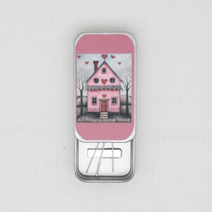 May include: A pink metal tin with a hinged lid, featuring a painted illustration of a pink house with a red door, hearts, and the text "All hearts come home." The tin contains silver paper clips.