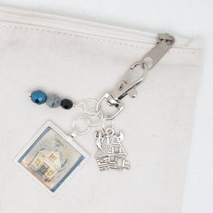 May include: A silver charm with a small house design, a silver charm with a picture of a house, and a silver charm with blue, gray, and black beads. The charms are attached to a silver key chain.