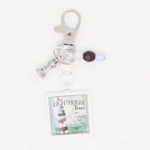 May include: A silver keychain with a small lighthouse charm, a brown and blue bead, and a square charm with a lighthouse image and the text "The Lighthouse Inn" and "We'll Keep The Light On For You".