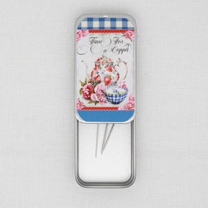 May include: A silver metal tin with a blue and white gingham lid. The lid features a colourful illustration of a teapot, a bowl, and flowers. The text "Time for a Cuppa" is printed on the lid. The tin is open and contains several sewing needles.