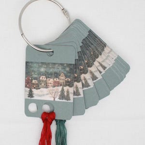 May include: A set of small, rectangular cards attached to a silver ring. Each card features a winter village scene with snow-covered houses and trees. Red and green threads are attached to the bottom of the first card.