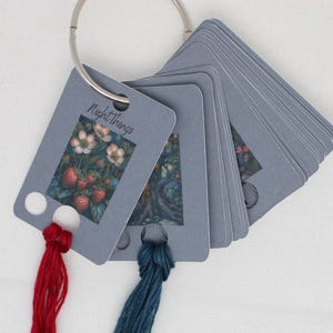 May include: A set of grey index cards on a silver ring, featuring botanical illustrations and the text "Night Things." Red and teal thread tassels hang from the cards. The cards display images of strawberries and flowers.