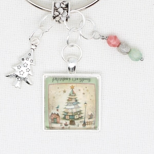 May include: A silver-toned Christmas-themed charm with a square pendant featuring a Christmas tree illustration. The charm includes a tree-shaped dangle and beaded accents in pink, silver, and green.