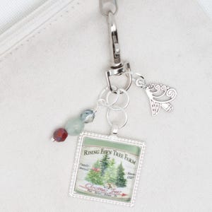 May include: Silver charm with a green and white image of a Christmas tree farm with the text "Rising Fawn Tree Farm" and "Christmas Trees". The charm is attached to a silver key chain with a silver butterfly charm and green and red beads.