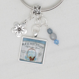 Puede incluir: Una pulsera de plata con un dije floral, cuentas azules y un colgante cuadrado. El colgante presenta una ilustración azul y blanca de una cesta de hortensias con el texto "Blue Mountain Hydrangea Farm".