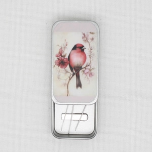 May include: A silver metal needle case with a floral design featuring a red bird. The case has a hinged lid and a compartment for storing needles.