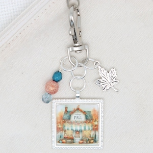 Op de afbeelding: Een zilverkleurige sleutelhanger met een vierkante hanger met een illustratie van een winkel versierd voor de herfst, inclusief de tekst FALL. De sleutelhanger bevat een zilveren esdoornbladbedel en drie kralen in blauwe en oranje tinten.