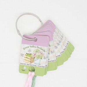 May include: A set of 10 fabric swatches with a pink and green color scheme. The swatches are attached to a silver ring and feature a label that reads "Green Valley Orchards" with a picture of apples and hydrangeas. The label also lists the apple varieties: Granny Smith, Rhode Island Greening, and Newtown Pippin.