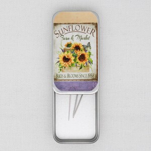 May include: A silver metal tin with a sunflower design and the text "Sunflower Farm & Market Buds & Blooms Since 1958". The tin is open and contains three sewing needles.