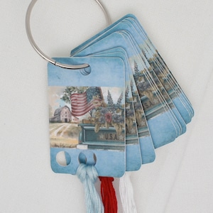 May include: A set of blue and white cards with a patriotic theme, featuring a red, white and blue American flag, a barn, and a truck with the words "U.S.A." on it. The cards are attached to a silver ring and have three strands of embroidery floss hanging from them.