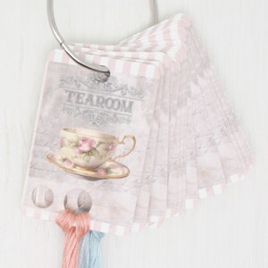 May include: A set of decorative tags with a metal ring. Each tag features a vintage design with a teacup illustration and the word "TEAROOM". The tags have a pink and white striped border and are attached to a metal ring. Pink and blue threads are attached to the tags.