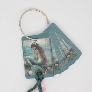 May include: A set of teal blue cards with a mermaid illustration on the front card. The cards are attached to a silver ring and have a green and pink tassel.