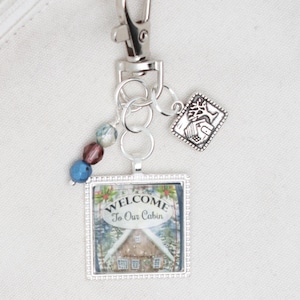 May include: Silver keychain with a square charm featuring a winter cabin scene and the text "Welcome to Our Cabin". The charm is attached to a silver chain with a silver split ring and a silver charm of a cabin with a tree. There are also three blue and brown glass beads on the chain.