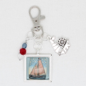 Pode incluir: Um chaveiro com tema náutico com um fecho mosquetão prateado. Apresenta um pingente quadrado com um desenho de veleiro, um pequeno berloque de veleiro e um berloque com contas vermelhas, brancas e azuis. O veleiro tem velas listradas em vermelho e branco.