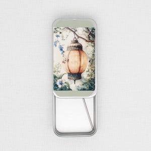May include: A silver metal tin with a hinged lid. The lid features a watercolour illustration of a hanging lantern with a floral design. The lantern is a light orange colour with a white and blue trim.