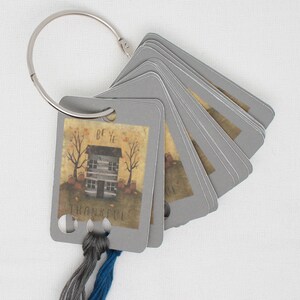 May include: A gray card holder with a ring at the top. The card holder has a small house illustration with the words "Be Ye Thankful" on it. There are two strands of embroidery floss threaded through the holes in the card holder.