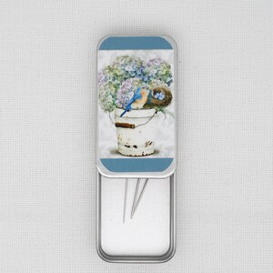 May include: A metal tin featuring a bluebird perched in a white bucket overflowing with blue and purple hydrangeas. The tin is open, revealing four silver sewing needles inside.