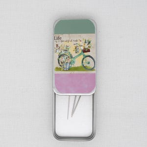 May include: A silver metal tin with a floral design and the text "Life is a beautiful ride". The tin is open and contains three silver sewing needles.