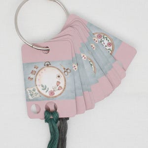 Puede incluir: Un conjunto de tarjetas de hilo de bordar rosa y azul con un diseño floral y un aro con herramientas de bordado. Las tarjetas están unidas a un anillo plateado.