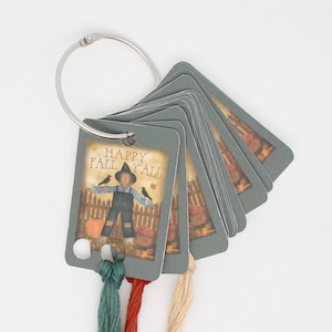 May include: A set of gray cards with a scarecrow illustration and the text "Happy Fall Y'all". The cards are attached to a silver ring and have four strands of embroidery floss in green, red, beige, and cream colors.
