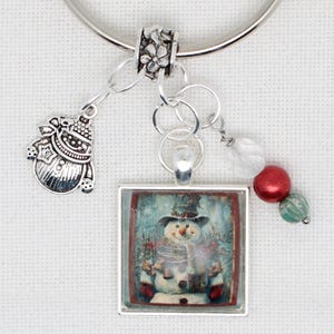 May include: A silver charm bracelet with a snowman pendant. The pendant features a square image of a snowman wearing a hat and scarf. Additional charms include a snowman-shaped charm and red and green beads.