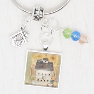 May include: A silver key chain with a charm of a birdhouse with a heart on the door. The charm is attached to a square glass pendant with a picture of a birdhouse with two birds on it. The key chain also has a silver bead with flowers on it and three glass beads in green, blue, and pink.