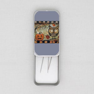 May include: A rectangular, silver needle case with a Halloween design. The lid shows a pumpkin, an owl, and autumn leaves. The case is open, displaying needles. The background is light blue.