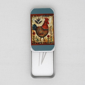 May include: A metal tin with a hinged lid. The lid features a colorful illustration of a rooster with a red comb and wattles. The tin is open and contains several sewing needles.
