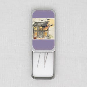 May include: An open, rectangular metal tin with a purple lid. The lid displays a Halloween scene: a house, a witch on a broom, bats, and a full moon. The tin contains needles. The overall design has a vintage feel.