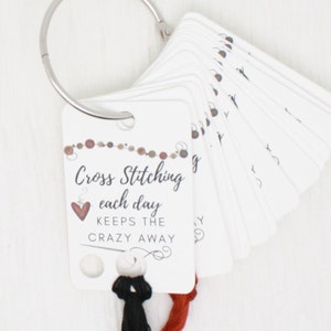 May include: A set of white cards on a silver ring, with the text "Cross Stitching each day keeps the crazy away." The cards have a heart and decorative elements. Below the cards are black and red threads.
