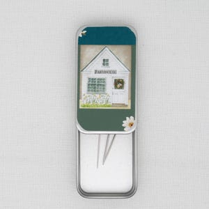 May include: A metal tin with a white farmhouse illustration on the lid. The tin is open and contains three silver needles.