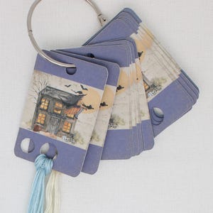 May include: A set of Halloween-themed tags with a house illustration, attached to a metal ring. The tags have a blue border and a hole for attaching thread. The illustration features a witch, bats, and a full moon.