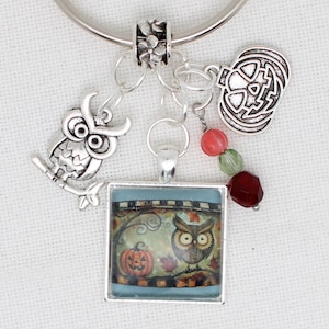 May include: Silver charm bracelet with Halloween-themed charms. Includes an owl on a branch, a jack-o'-lantern, and a square pendant with an owl and pumpkin scene. The bracelet also features colourful beads.