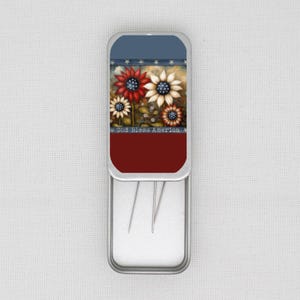 May include: A rectangular metal needle case with a patriotic design. The top section is blue and red, with a floral design and the text "God Bless America". The case is open, revealing several needles.