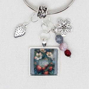 May include: A silver charm bracelet with a square pendant featuring a strawberry painting. The bracelet includes a strawberry charm, a flower charm, and decorative beads in shades of grey, pink, and red. The bracelet is on a white background.