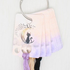 May include: A set of colourful embroidery floss cards with a Halloween theme. The cards feature a black cat sitting on a crescent moon with bats flying around it. The cards are held together with a silver ring.